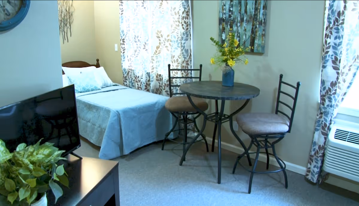 A cozy senior living room featuring a small round table with two cushioned chairs, a bed with light blue bedding, a TV on a stand, a potted plant, floral patterned curtains, and a wall-mounted air conditioning unit.