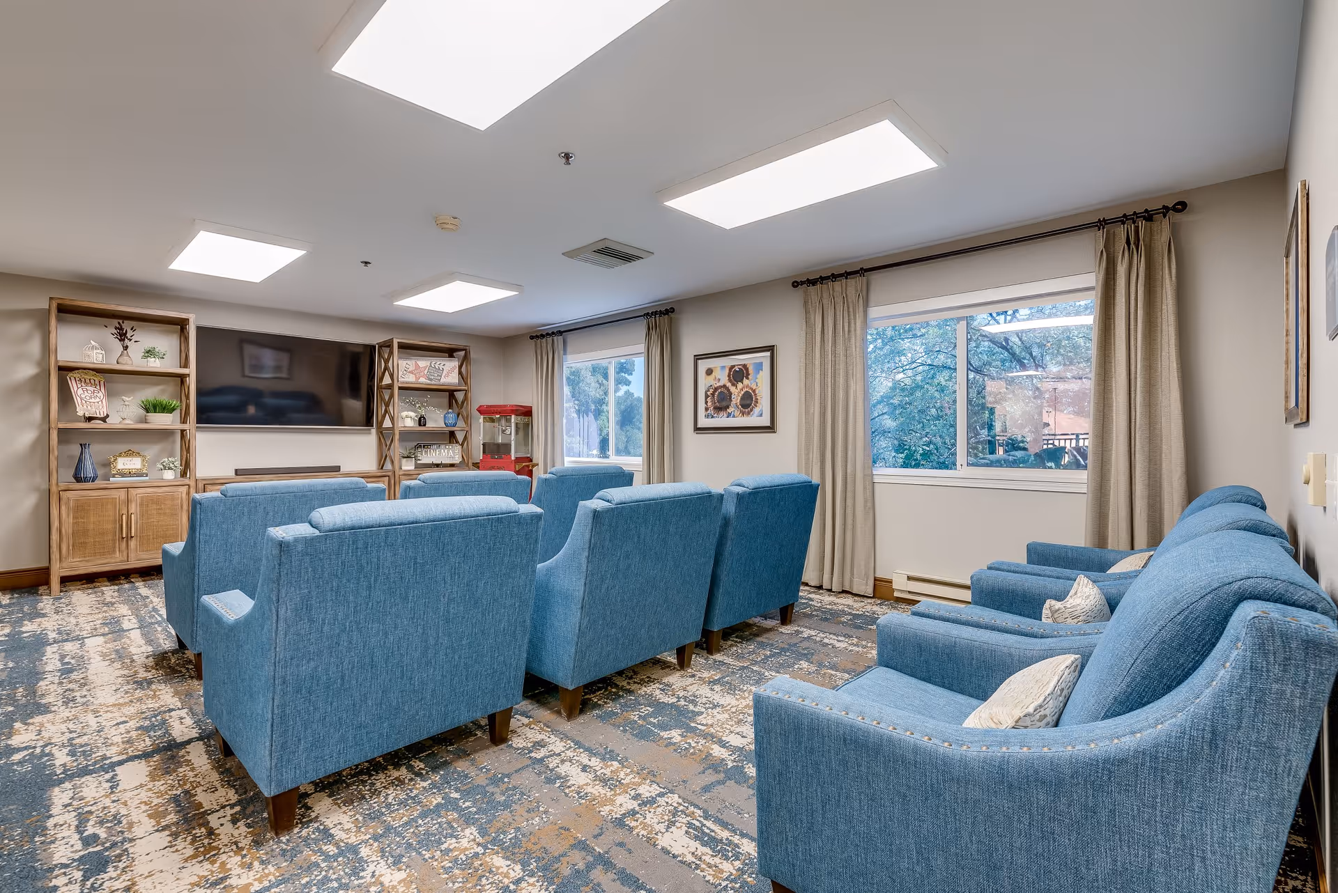 A cozy senior living common area with multiple blue upholstered armchairs arranged in rows facing a large flat-screen TV mounted on the wall. The room features two large windows with beige curtains, wooden shelving units with decorative items, and a patterned carpet. The ceiling has recessed lighting panels providing bright illumination.