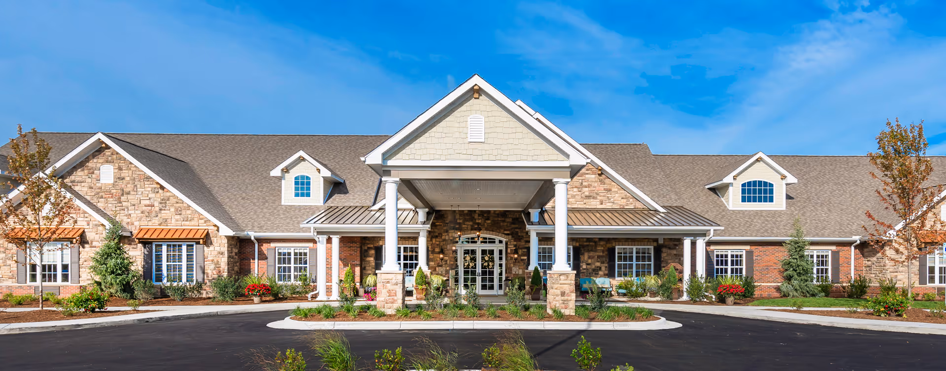 Front exterior view of a single-story senior living facility building with a covered entrance supported by white columns, stone and brick facade, multiple windows, and a clear blue sky above.
