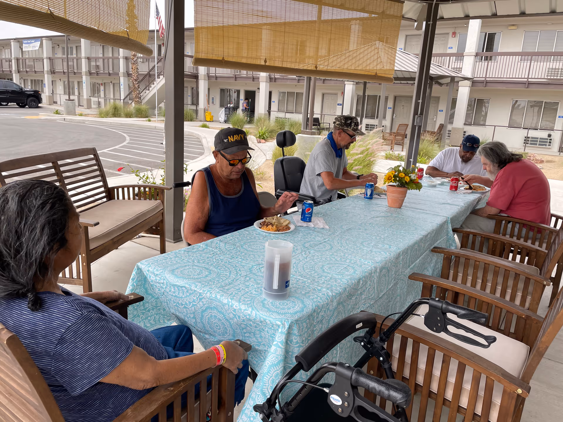 Five elderly individuals sitting around a rectangular table covered with a light blue patterned tablecloth under a shaded outdoor patio. They are eating and drinking soda cans. The background shows a multi-story building with balconies and a parking area.