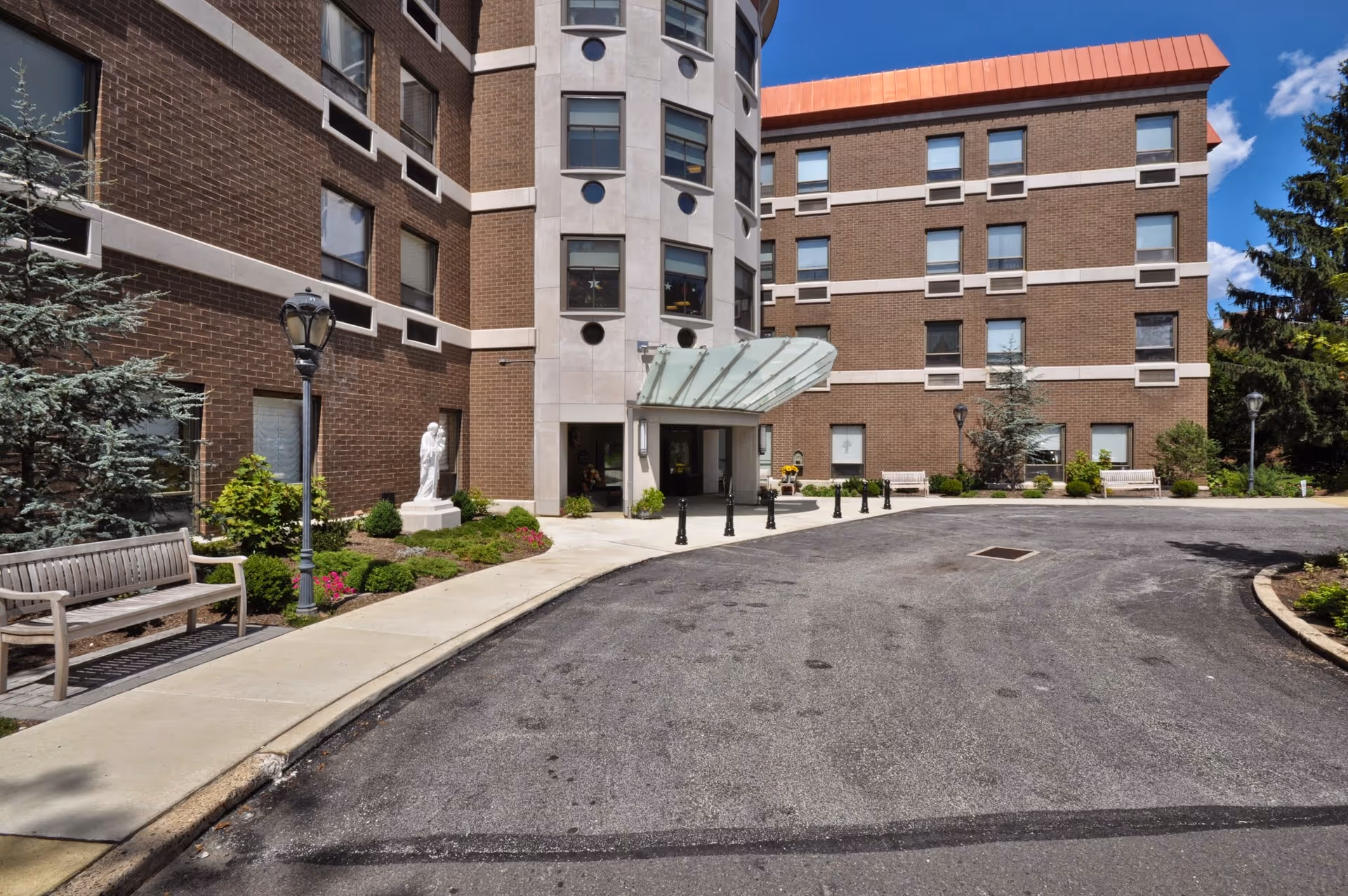 Front entrance of a multi-story brick building with a glass canopy, benches, statue, landscaping, and a circular driveway.