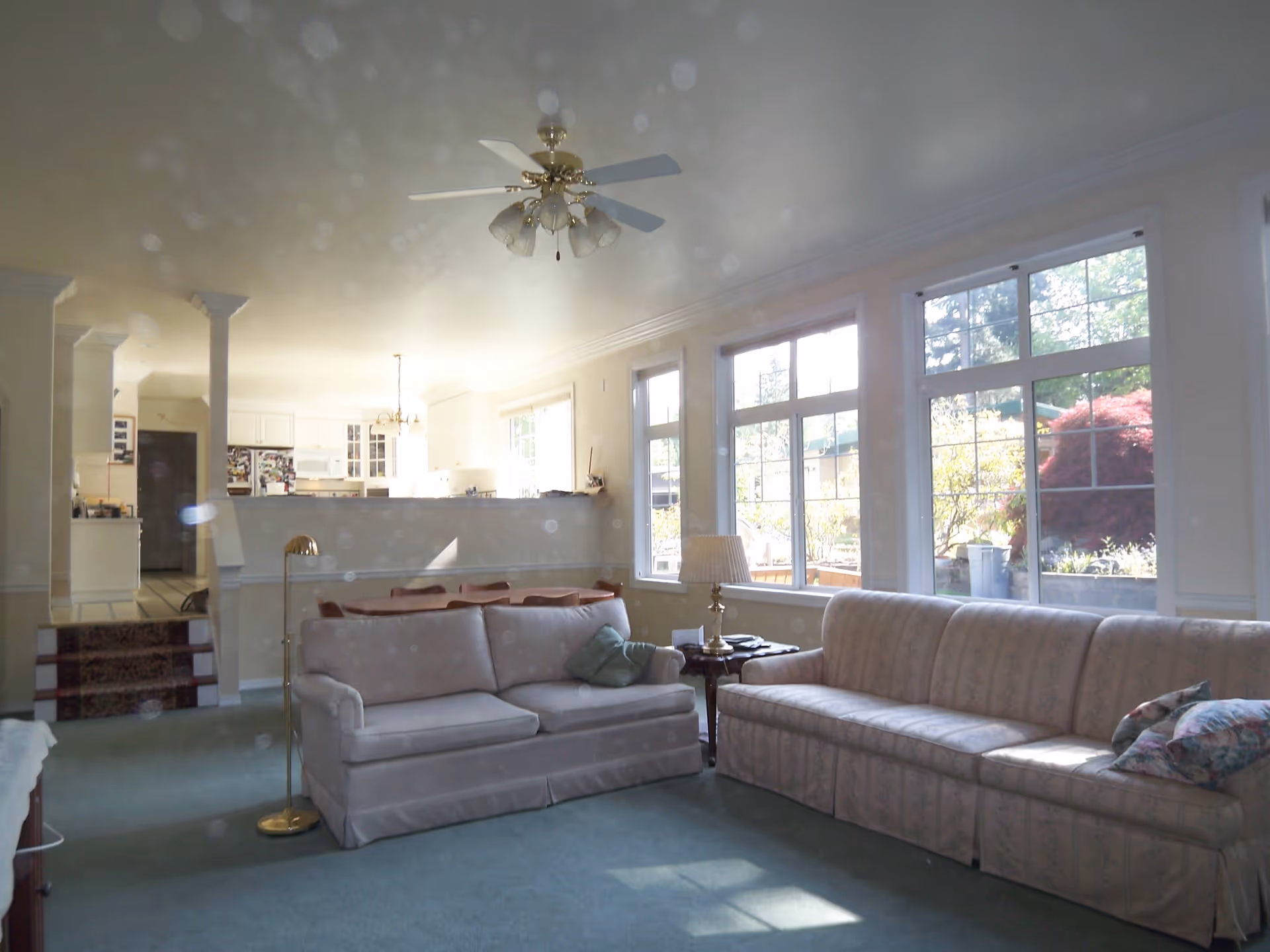 A bright living room with two beige sofas, a small wooden side table with a lamp, large windows letting in natural light, and a ceiling fan. In the background, there is a dining area with a wooden dining table and chairs, and a kitchen with white cabinets and appliances.