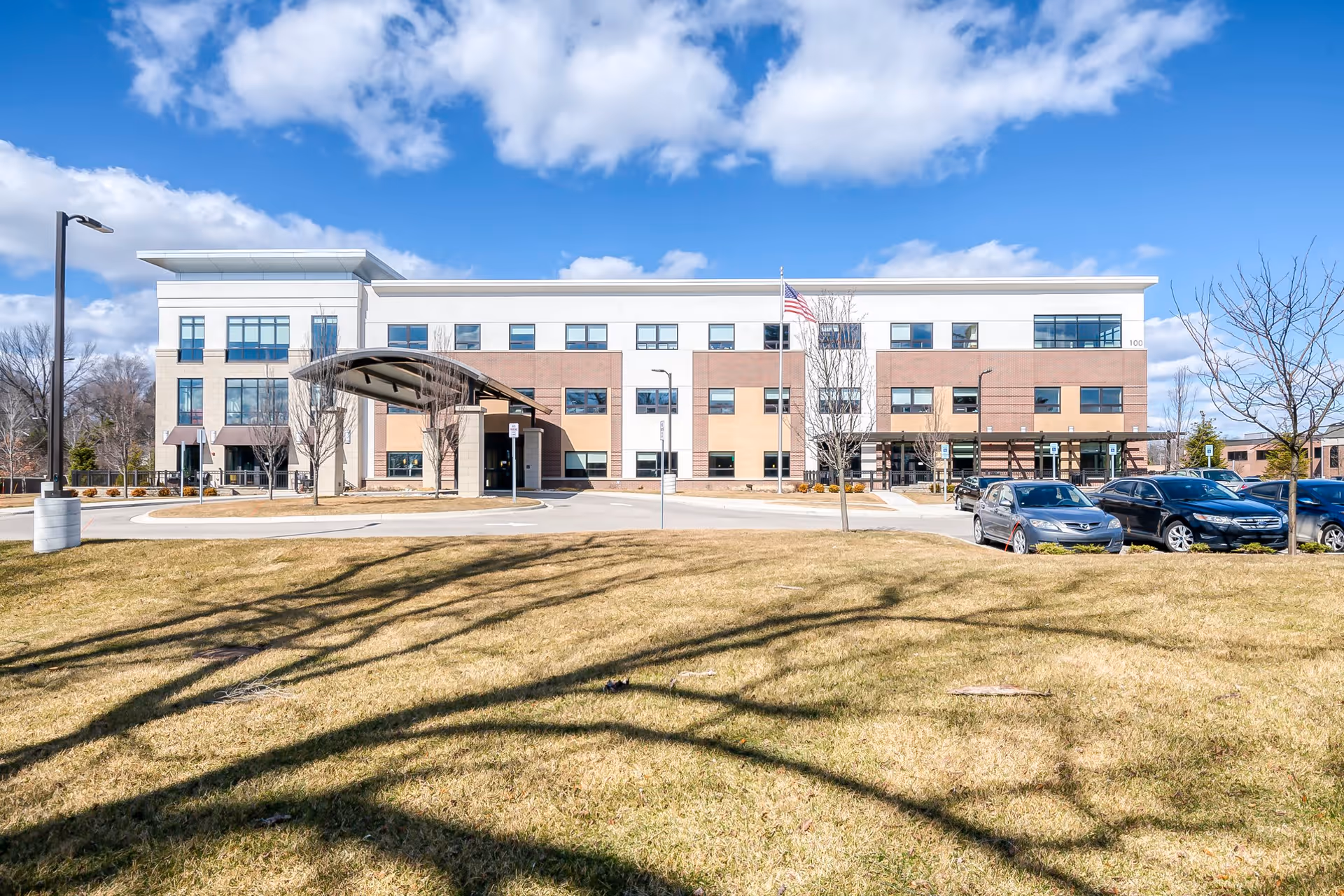 Front exterior view of The Avalon of Bloomfield Township, a modern three-story building with a covered entrance, several windows, an American flag, parked cars, and a grassy area with leafless trees in the foreground under a partly cloudy blue sky.