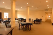 A spacious dining room with multiple dark wooden tables and chairs arranged evenly across a beige carpeted floor. The room has cream-colored walls, several columns, recessed ceiling lights, and a long hallway visible on the right side.