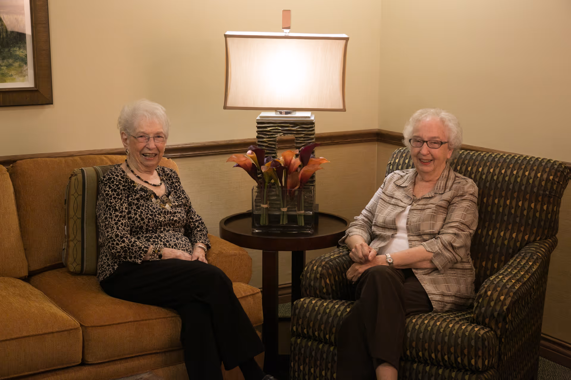 Two elderly women sitting and smiling in a cozy living room area with a lamp and flower arrangement on a small round table between them.