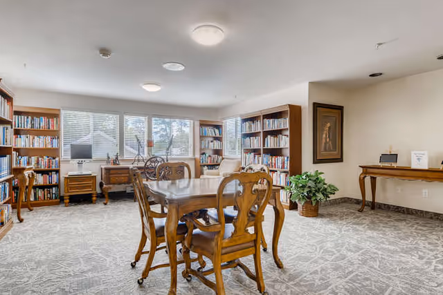 A bright and spacious room with large windows letting in natural light. The room features wooden bookshelves filled with books along the walls, a wooden table with four matching chairs in the center, and a wooden desk with a computer and decorative items near the windows. There is a framed painting on the wall and a potted plant on the floor. The carpet has a subtle patterned design.