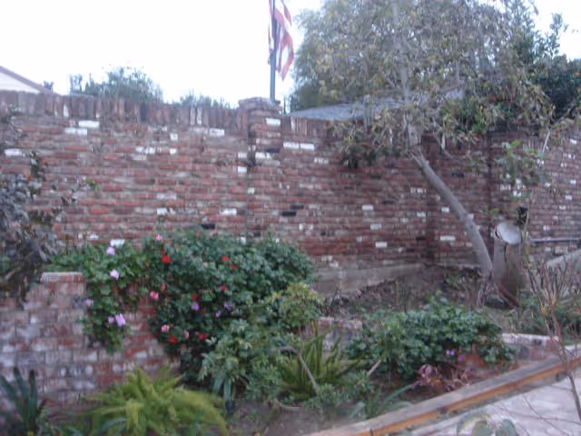 Brick courtyard with flowering plants, a garden, and a flag atop a weathered brick wall.