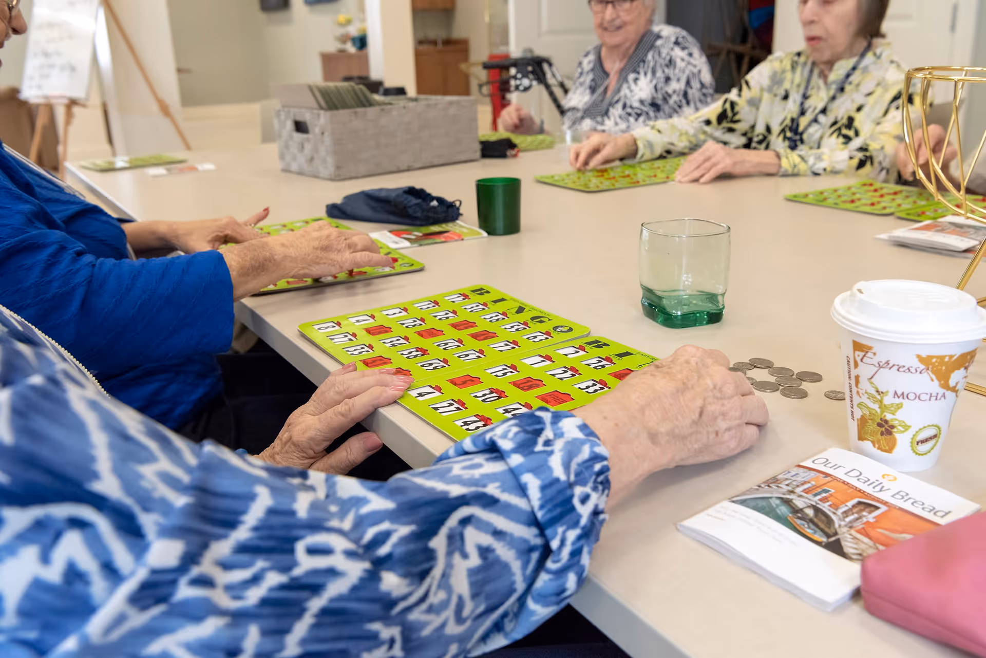 A group of elderly individuals sitting around a table playing bingo. The table has bingo cards, coins, a cup of coffee, a glass, and a newsletter titled 'Our Daily Bread'.