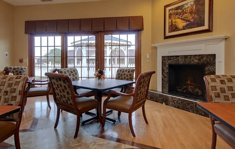 A cozy dining area with a square table surrounded by four cushioned chairs with patterned upholstery. Behind the table, large glass doors reveal an outdoor gazebo and white picket fence. To the right, there is a fireplace with a dark marble surround and a framed painting above it. The room has warm lighting and wooden flooring.