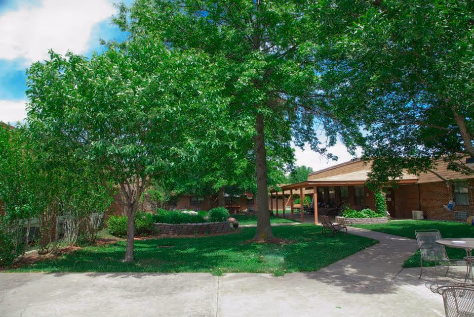 Outdoor courtyard area with green grass, several trees, and a paved walkway. There are patio chairs and tables on the right side, and a covered porch attached to a brick building in the background under a partly cloudy blue sky.