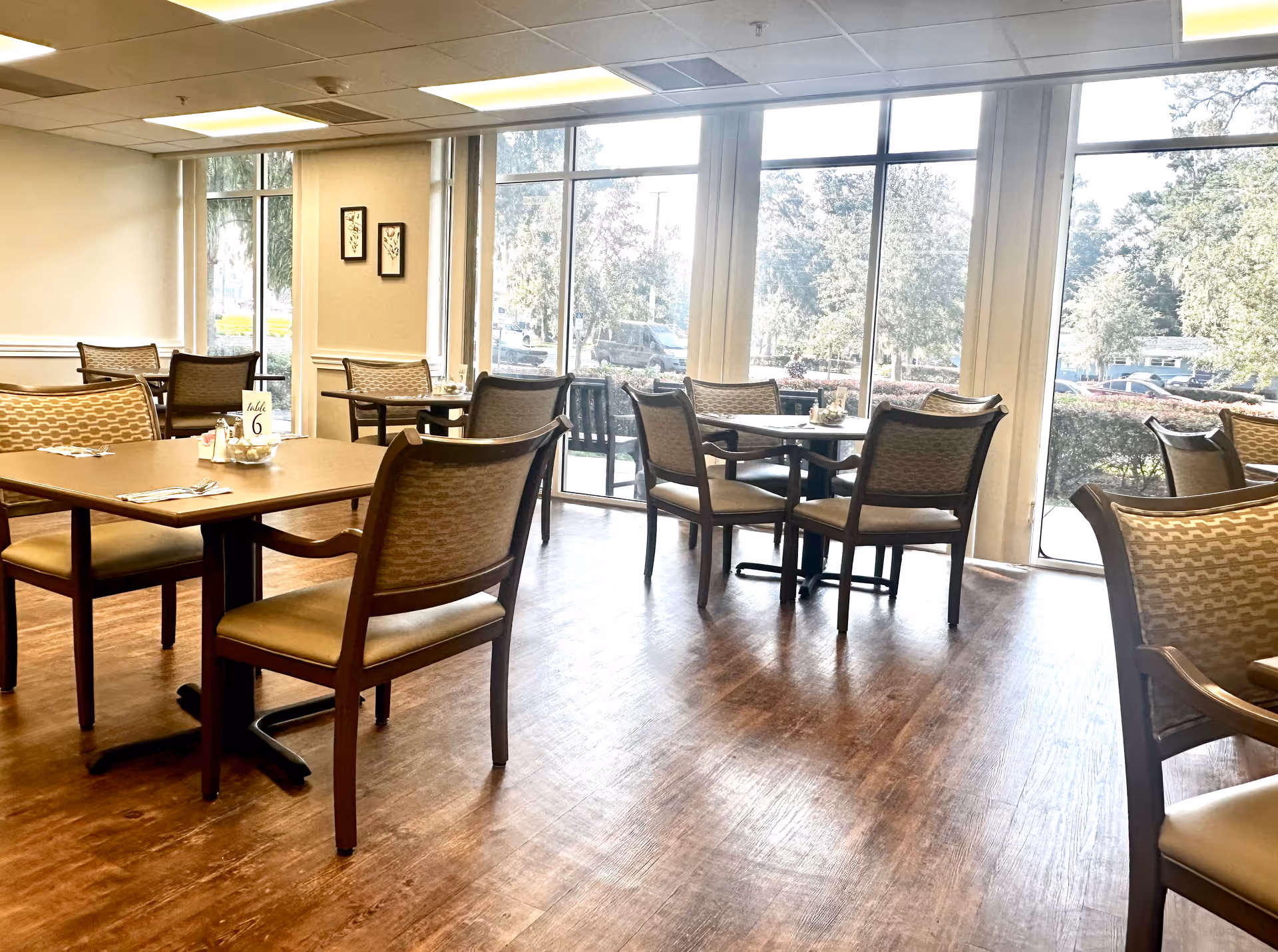 A bright dining room with several tables and chairs arranged neatly. Large windows allow natural light to fill the space, offering a view of trees and parked cars outside. The floor is wooden, and the walls are light-colored with minimal decorations.