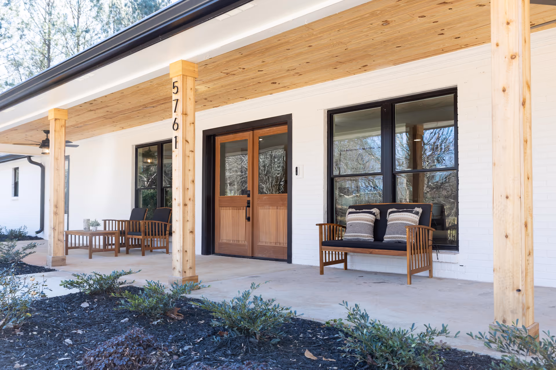 Front porch of a senior living facility with wooden pillars, a wooden double door entrance, black-framed windows, and outdoor seating including a bench with two pillows and two chairs with a small table. The porch ceiling is wooden, and there are plants and mulch in the garden bed in front.