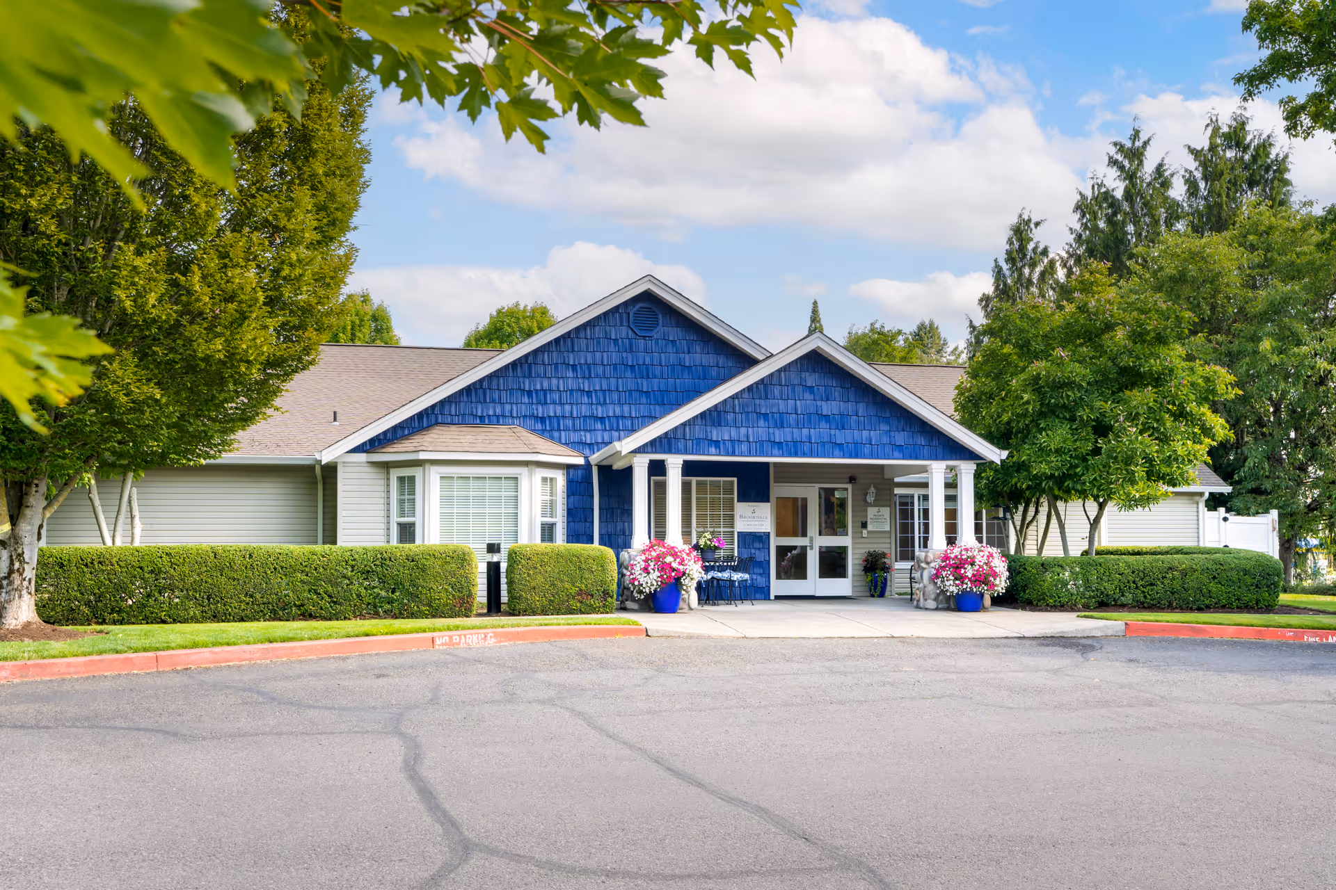 Front exterior view of a single-story building with blue siding and white trim, surrounded by green trees and bushes, with a paved driveway and flower pots near the entrance under a partly cloudy sky.