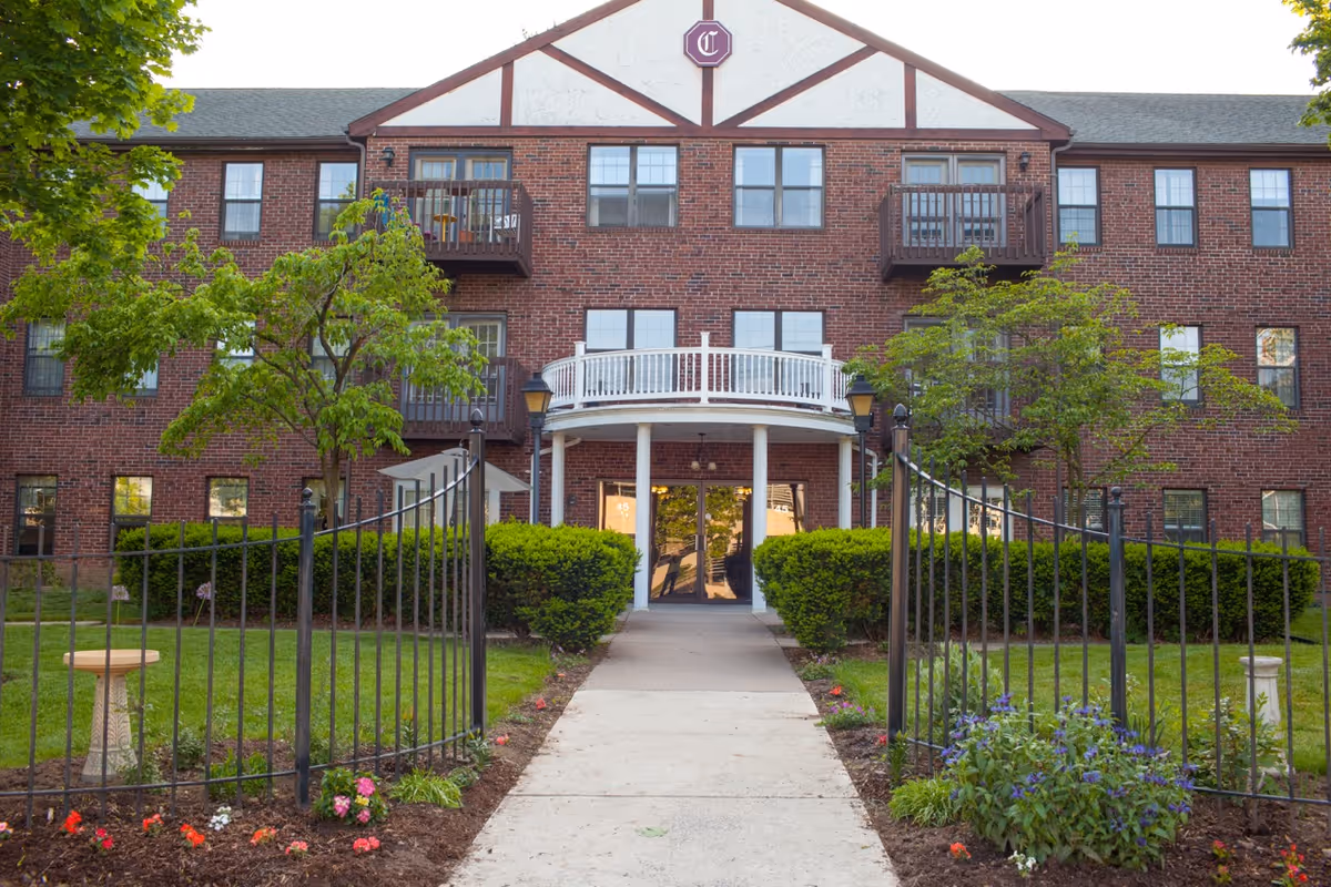Front exterior view of a three-story brick building with a white balcony above the entrance, surrounded by green trees, bushes, and a black metal fence with a concrete walkway leading to the entrance.