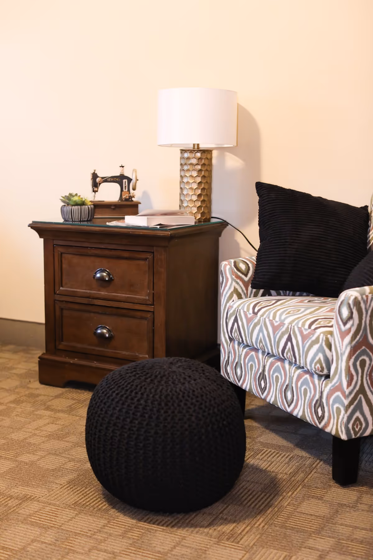 A cozy corner featuring a patterned armchair with black cushions, a dark wooden side table with two drawers, a decorative lamp with a honeycomb-patterned base and white shade, a small potted succulent, a vintage sewing machine decor piece, and a black knitted pouf on a carpeted floor.