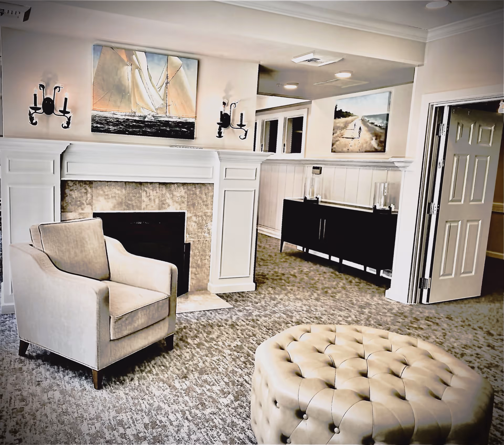 A cozy living room area featuring a beige armchair and a large tufted ottoman on a patterned carpet. The room has a white fireplace with a painting of a sailboat above it, flanked by two wall sconces. In the background, there is a black sideboard with decorative glass items and another painting depicting a beach scene. An open door is visible on the right side.