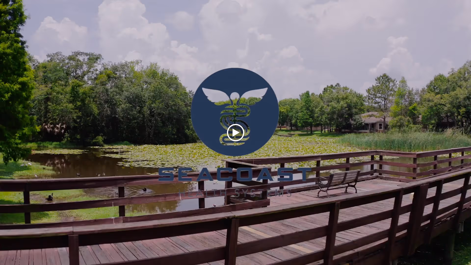 A wooden boardwalk with benches overlooking a pond covered with lily pads, surrounded by green trees under a partly cloudy sky.