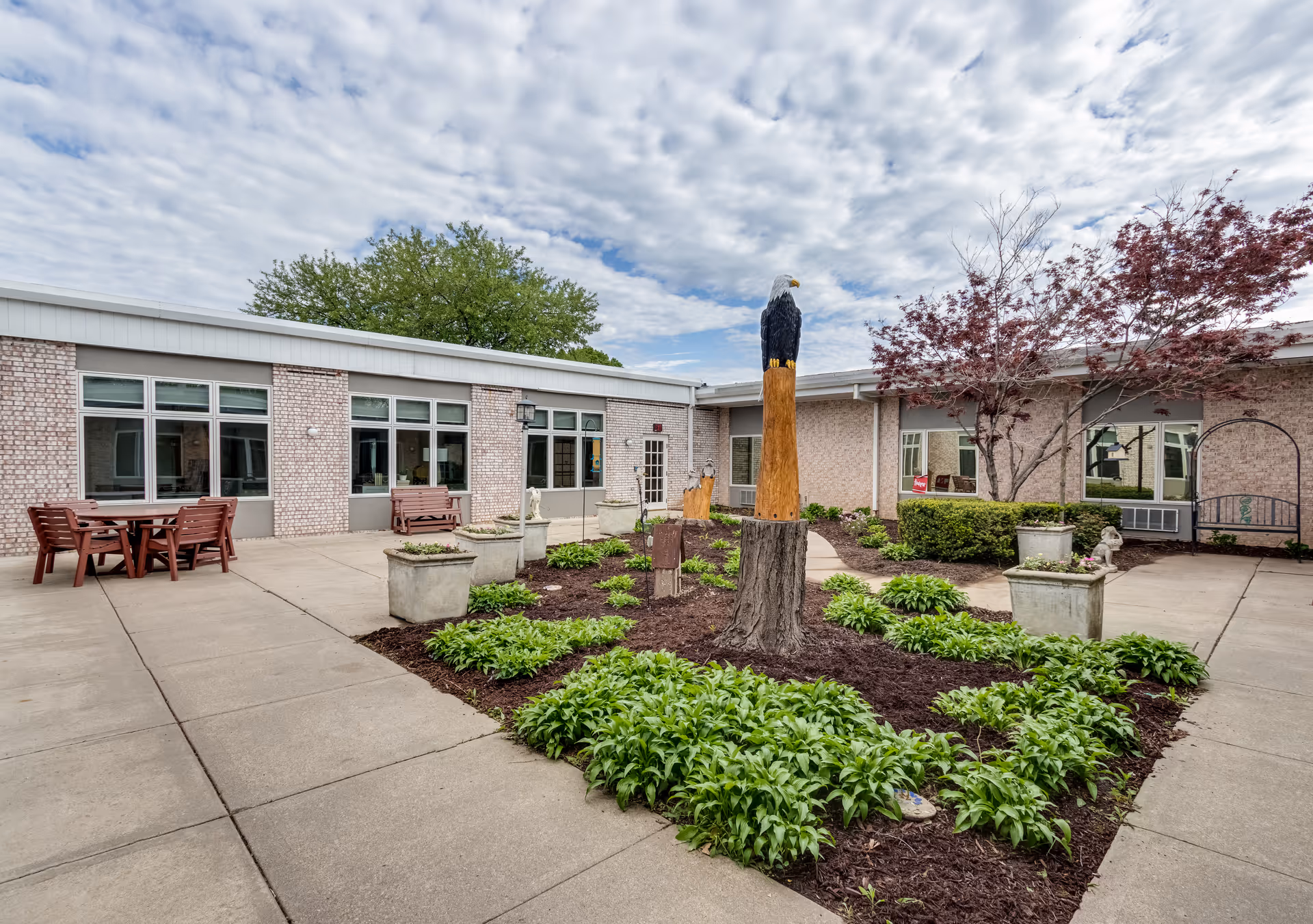 Outdoor courtyard area at Brookestone of Papillion featuring a landscaped garden with green plants and a carved wooden eagle statue on a tree stump. Surrounding the garden are concrete walkways, outdoor seating with tables and chairs, and a brick building with multiple windows. The sky is partly cloudy.