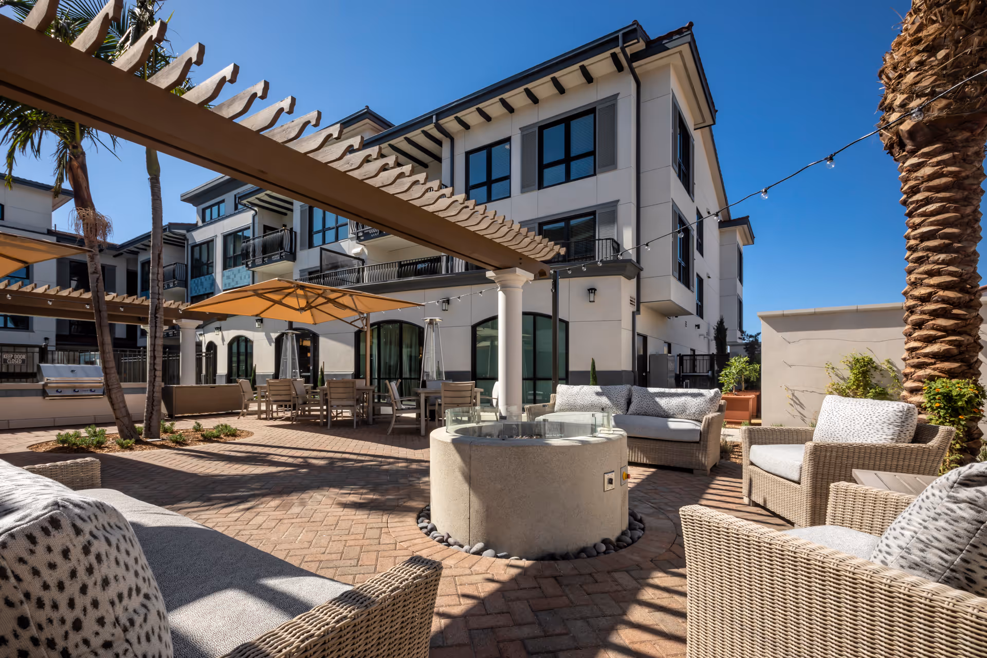 Outdoor patio area at Westmont of Cypress featuring wicker seating with cushions arranged around a circular fire pit, a pergola, dining tables with umbrellas, palm trees, and a multi-story building in the background under a clear blue sky.