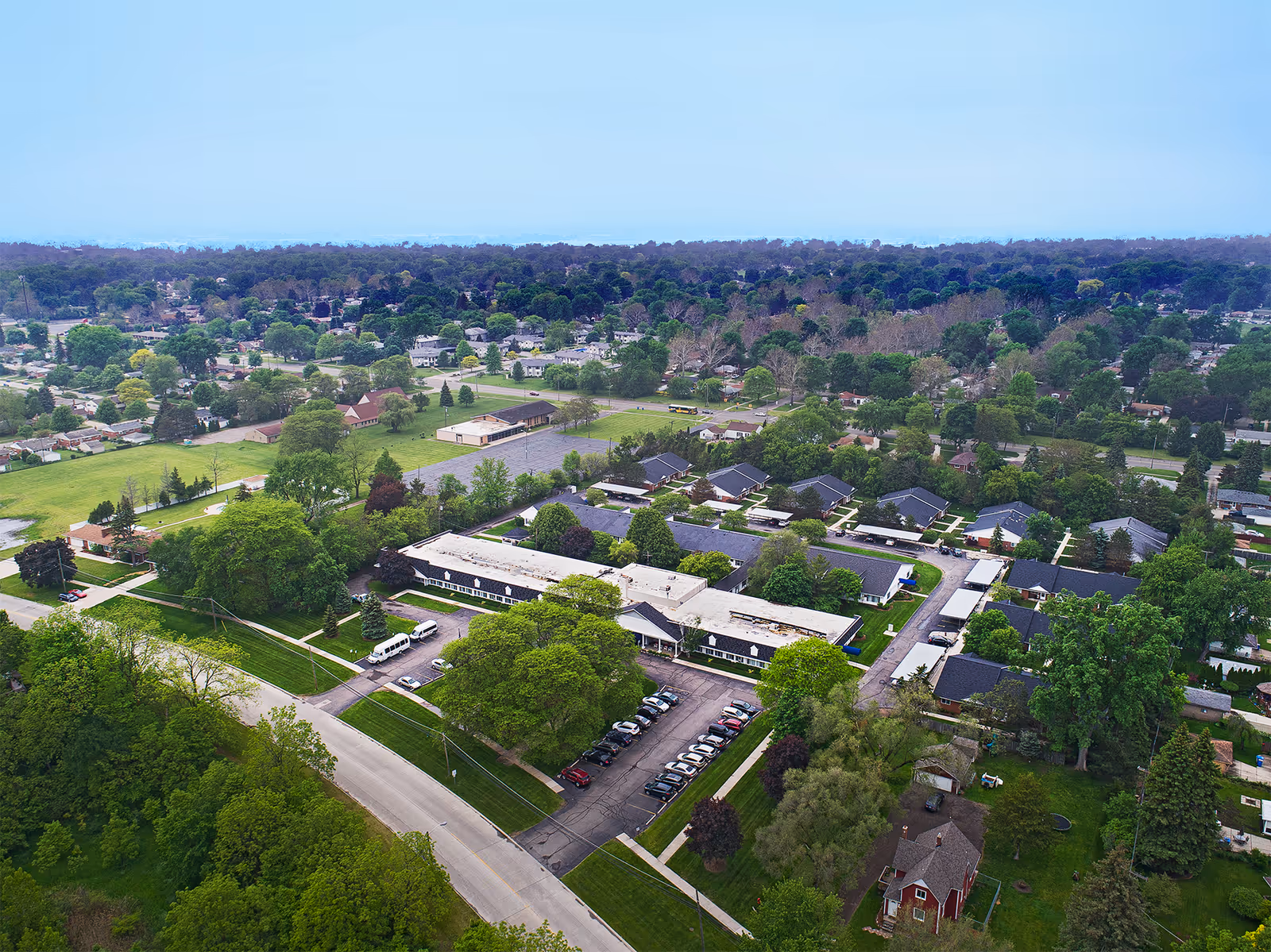 Aerial view of American House Dearborn Heights facility surrounded by trees and residential houses, showing multiple buildings, parking lots with cars, and green lawns under a clear sky.