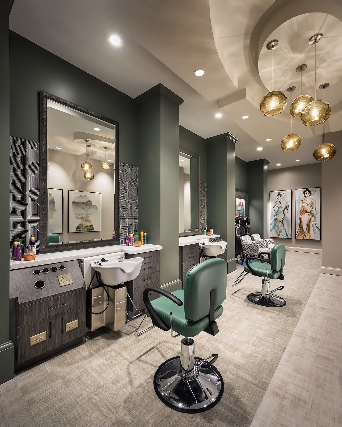 Interior of a modern hair salon with green walls and two green salon chairs in front of large mirrors. Each station has a white sink and shelves with hair care products. The ceiling features decorative hanging lights, and there are two framed fashion illustrations on the far wall.