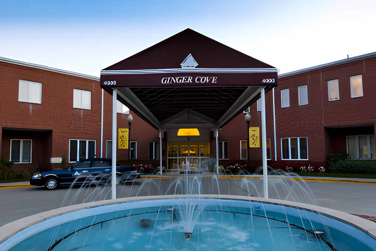 Entrance of Ginger Cove senior living facility with a maroon canopy over the drop-off, a fountain in the foreground, and a red brick building.