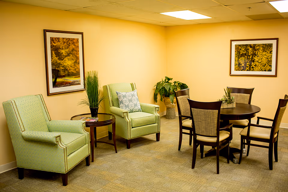 A cozy interior room with two light green upholstered armchairs separated by a small round wooden table with a plant on it. To the right, there is a round wooden dining table surrounded by six chairs with beige cushions. The walls are painted light yellow and decorated with framed pictures of autumn trees. A potted plant is placed in the corner of the room.