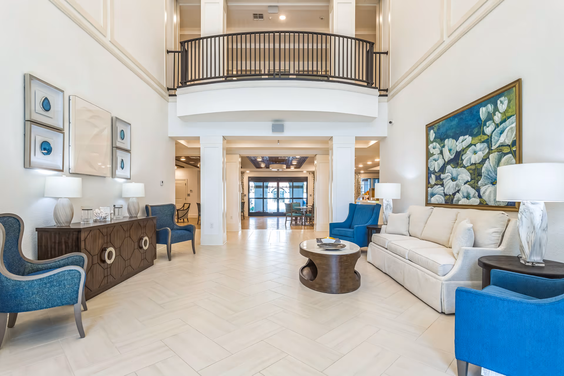 Bright and spacious living room area with high ceilings and a balcony railing above. The room features a beige sofa with white pillows, two blue armchairs, a round wooden coffee table, and side tables with white lamps. On the left wall, there is a wooden console table with two lamps and framed artwork above it. On the right wall, there is a large painting of white flowers on a blue and green background. The floor is light-colored tile arranged in a herringbone pattern. In the background, there is a dining area and glass doors leading outside.