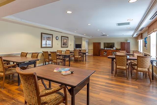 A spacious common room with several wooden tables and cushioned chairs arranged neatly on a wood floor. The room has light-colored walls with framed pictures and a clock. At the far end, there is a large flat-screen TV on a wooden cabinet with additional storage. Large windows on the right side let in natural light, and the ceiling has recessed lighting.