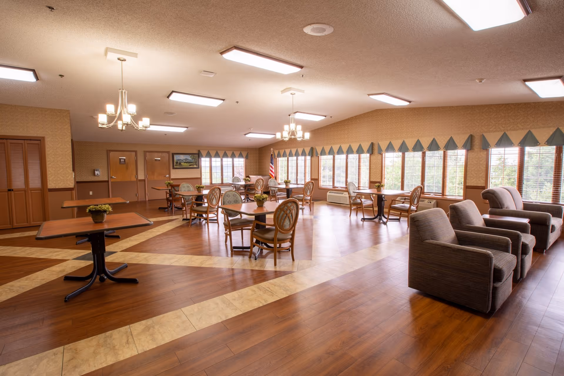 Spacious senior living dining/activity room with tables, chairs, armchairs, large windows, and chandeliers.