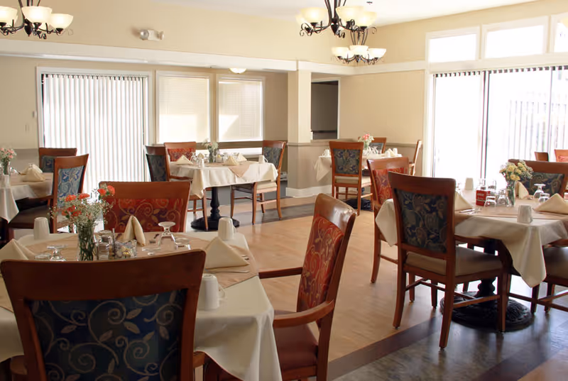 A dining room with multiple tables covered in white tablecloths, each set with napkins, glasses, and small floral centerpieces. The room has wooden chairs with patterned upholstery, large windows with vertical blinds, and two chandeliers hanging from the ceiling providing warm lighting.