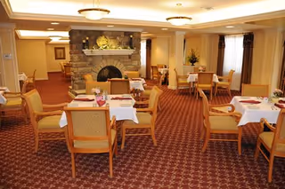 A cozy dining room with multiple tables covered with white tablecloths and set with plates and napkins. The room features a stone fireplace in the background, warm lighting, and carpeted floors with a red and beige pattern. Chairs with wooden frames and cushioned seats surround the tables, and windows with curtains allow natural light to enter.