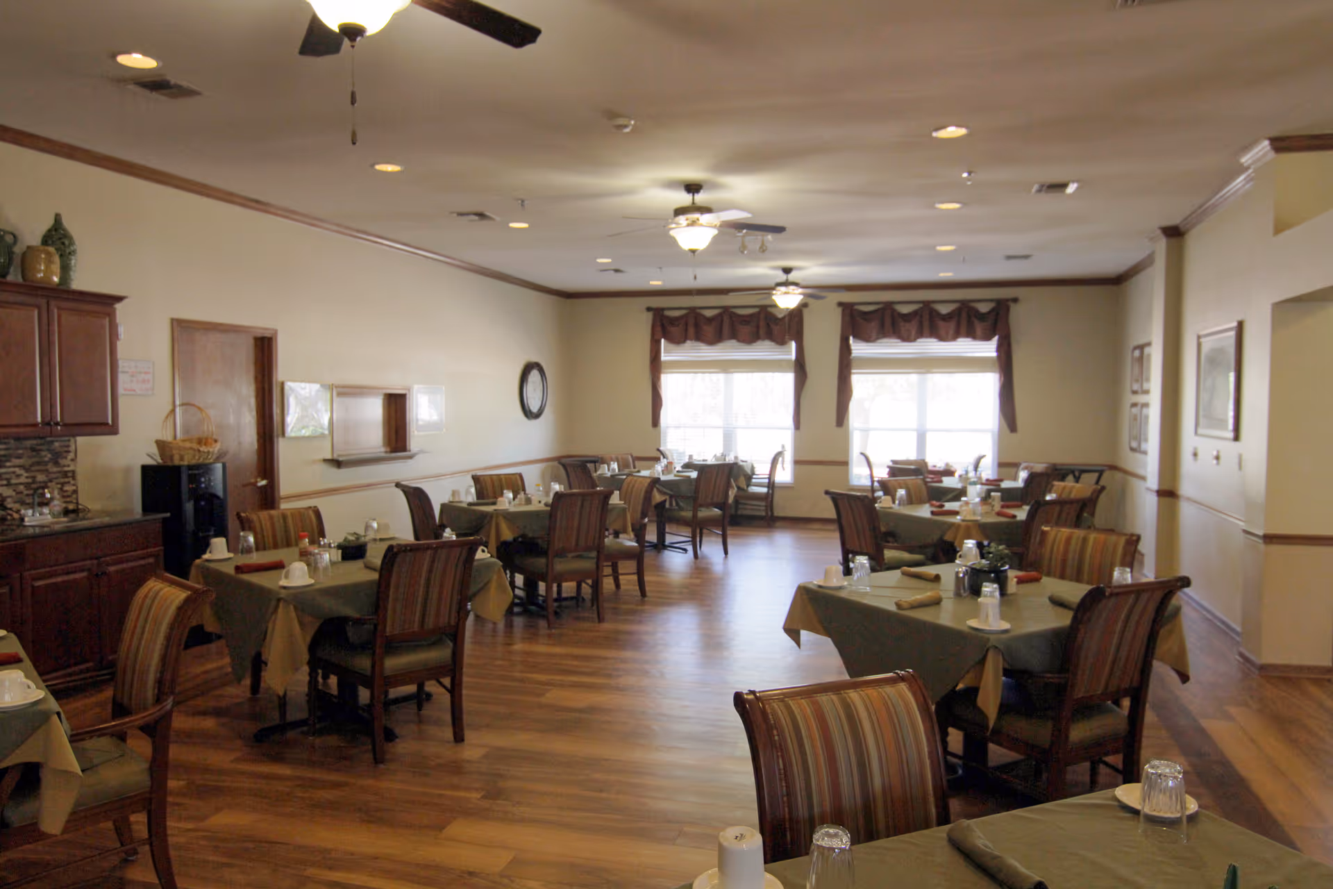 A dining room in a senior living facility with multiple tables covered in green tablecloths, each set with cups, glasses, and napkins. The room has wooden flooring, ceiling fans with lights, large windows with brown valances, and a wooden cabinet with decorative items on the left side.