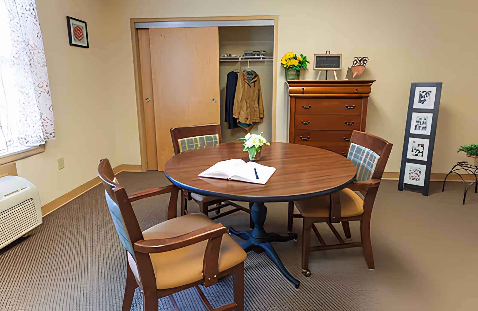 A cozy room with a round wooden table surrounded by four cushioned chairs. On the table, there is an open book with a pen and a small vase with white flowers. Behind the table, there is a wooden dresser with a small chalkboard, a vase with yellow flowers, and decorative items on top. To the left, there is a closet with sliding wooden doors, one partially open showing hanging clothes. A window with lace curtains lets in natural light, and a framed picture hangs on the wall. The room has beige walls and carpeted floor.