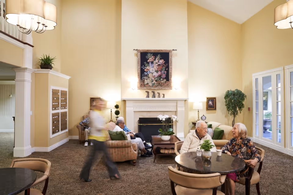 A cozy senior living common area with beige walls and carpeted floor. There are several elderly people seated on sofas and chairs around a fireplace with a floral tapestry above it. A woman is walking past in a blur. The room is decorated with plants, lamps, and framed pictures, and has large windows letting in natural light.