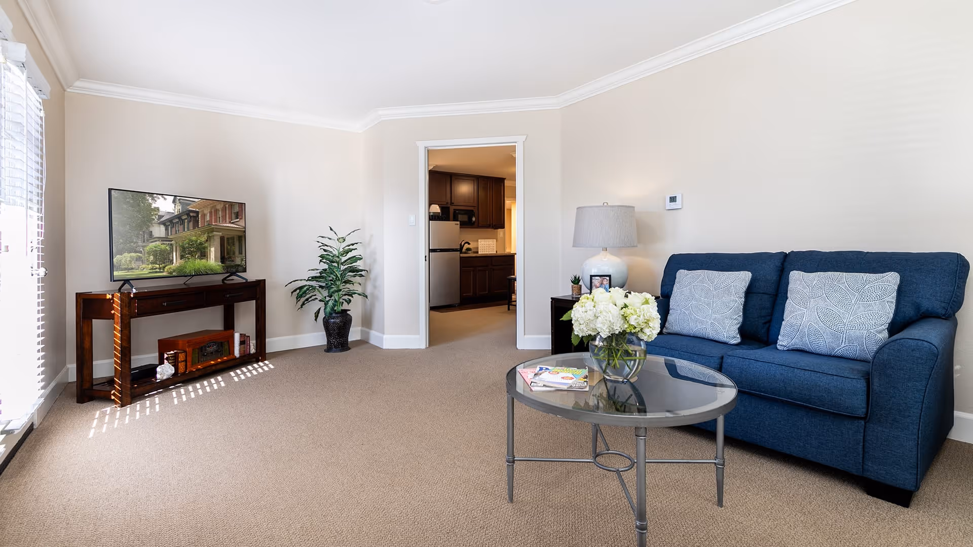 A bright living room with beige walls and carpet, featuring a blue sofa with two patterned cushions, a round glass coffee table with a vase of white flowers and magazines, a side table with a lamp and photo frame, a flat-screen TV on a wooden stand, and a potted plant near the doorway leading to a kitchen area.