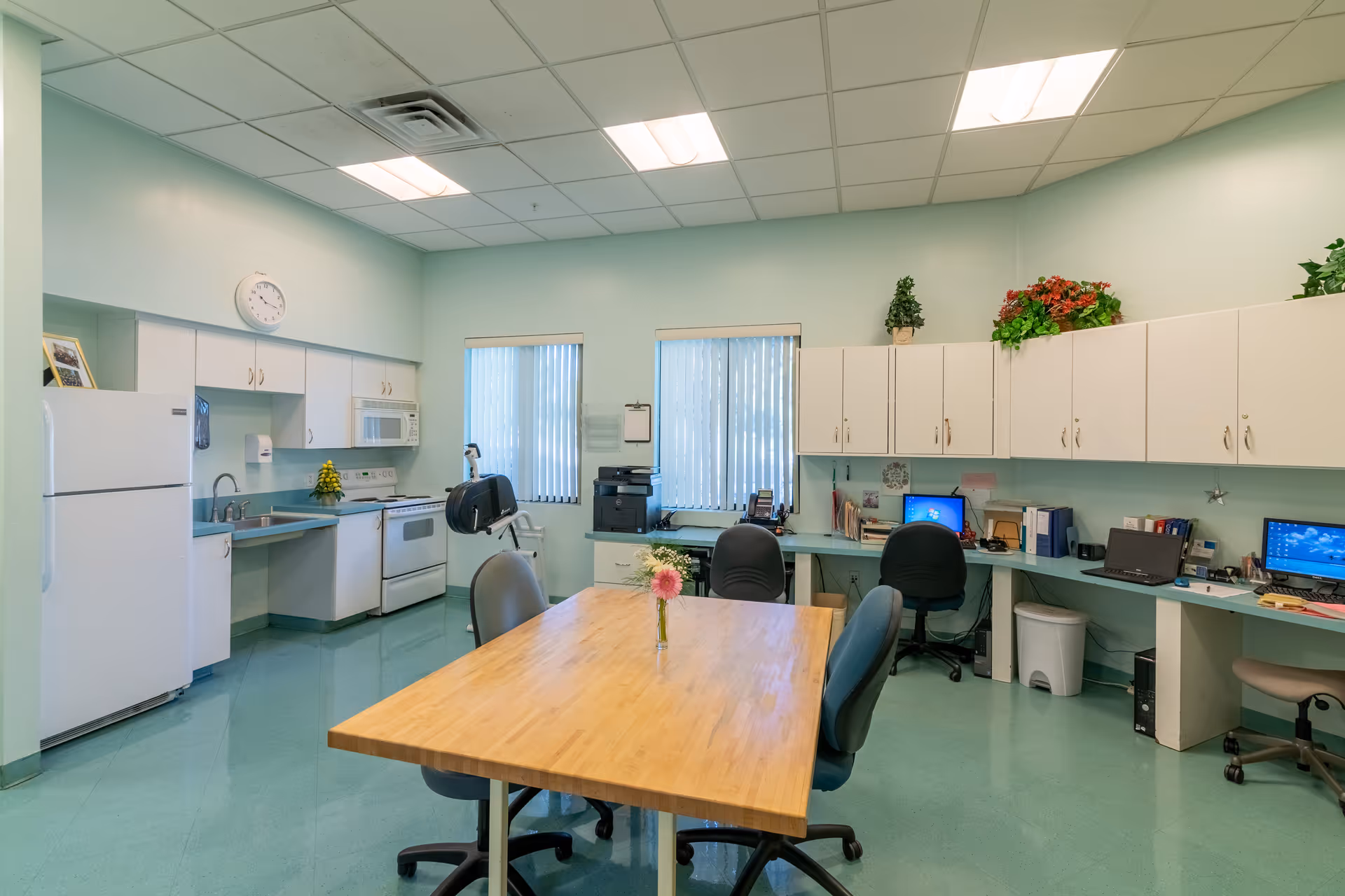 A bright, clean room with a kitchenette area including a refrigerator, microwave, stove, and sink on the left side. In the center is a wooden table with four office chairs around it. On the right side, there is a long desk with three computer workstations, office chairs, and cabinets above. The walls are light green, and there are two windows with vertical blinds. Decorative plants are placed on top of the cabinets.