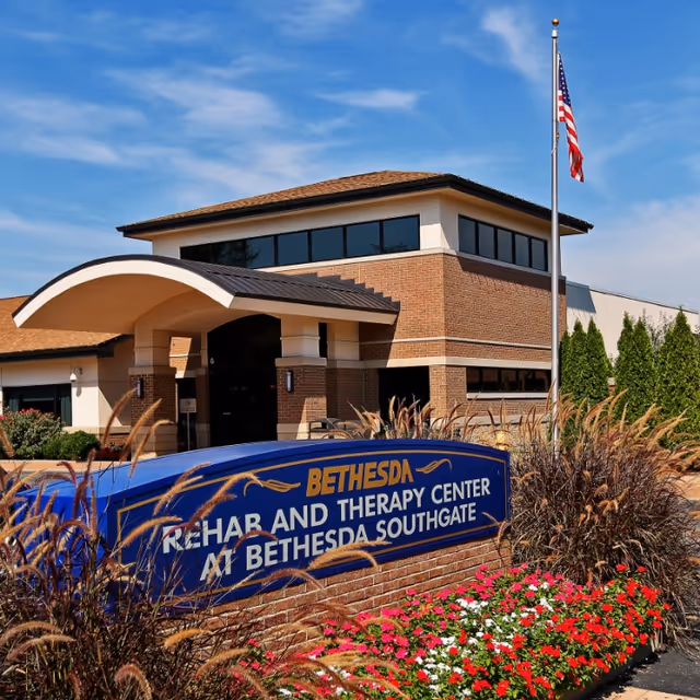 Exterior view of Bethesda Rehab and Therapy Center at Bethesda Southgate, showing a brick building with large windows, an arched entrance, an American flag on a flagpole, and a blue sign with yellow and white text surrounded by ornamental grasses and colorful flowers.