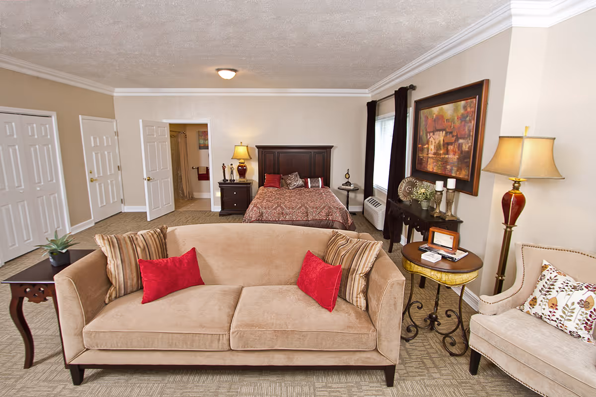 A cozy senior living bedroom featuring a beige sofa with red and striped pillows in the foreground, a bed with a patterned bedspread and multiple pillows in the background, a nightstand with a lamp and decorative items, a window with dark curtains, a side table with books and decor, an armchair with a floral pillow, and a floor lamp. The room has beige walls, carpeted floor, and a door leading to a bathroom.