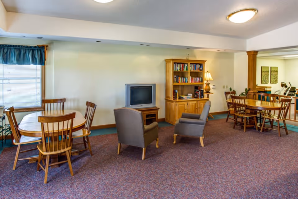 A senior living common room with round wooden dining tables and chairs, two armchairs facing a TV and a bookshelf.