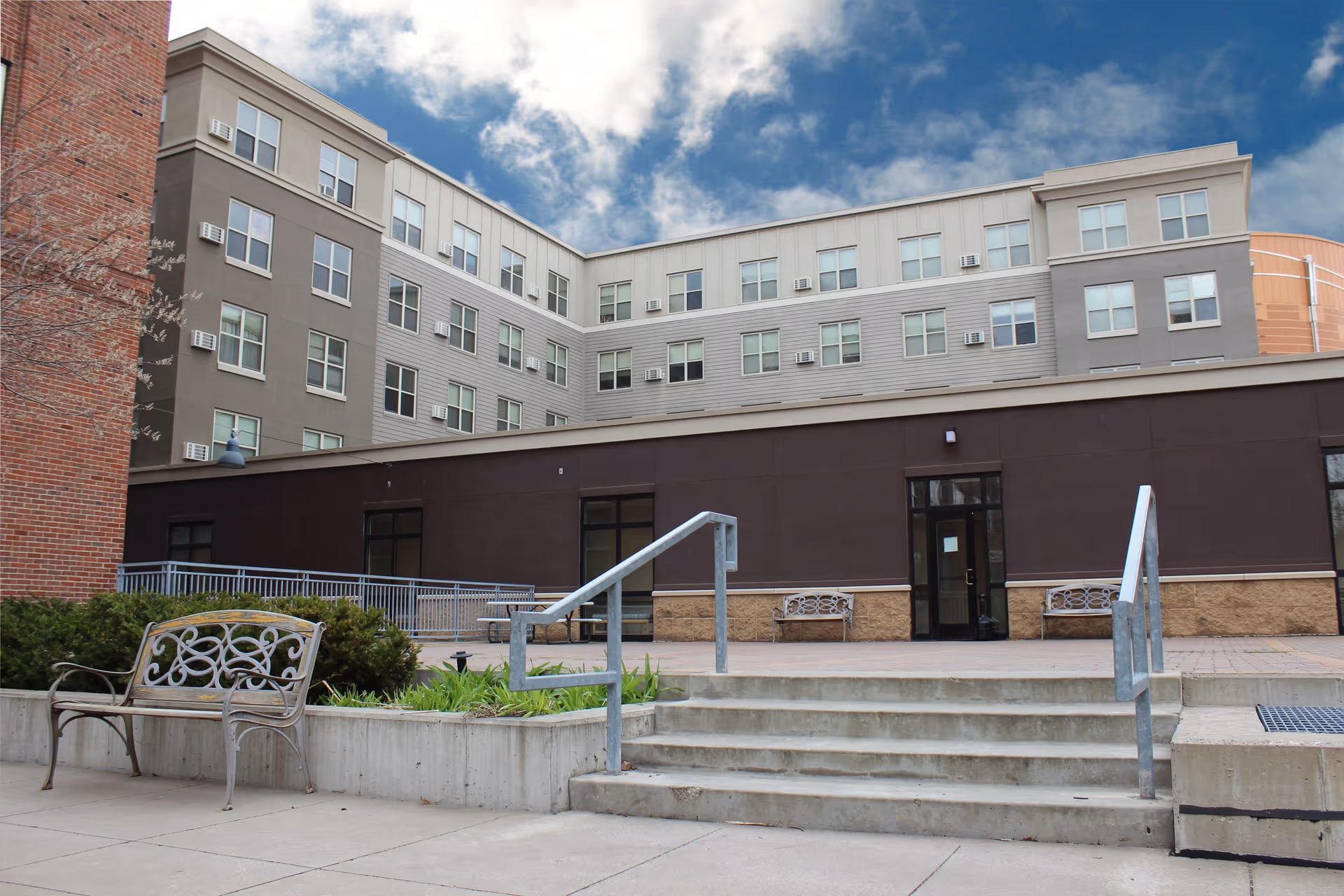 Exterior view of a multi-story senior apartment building with a courtyard area featuring benches, stairs with metal railings, and some greenery under a partly cloudy sky.