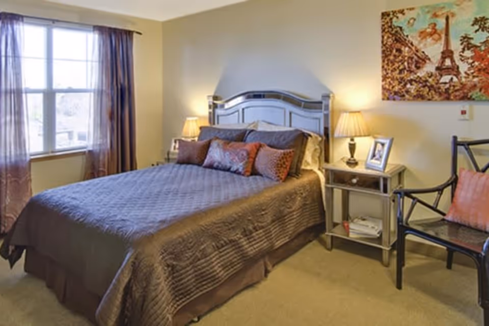 Well-lit bedroom with a bed dressed in brown bedding, a nightstand with a lamp and framed photo, a chair, window with curtains, and wall art.