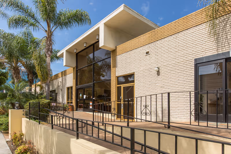 Exterior view of a single-story building with beige brick walls and large glass windows. There is a ramp with black metal railings leading to the entrance, surrounded by palm trees and other greenery under a clear blue sky.