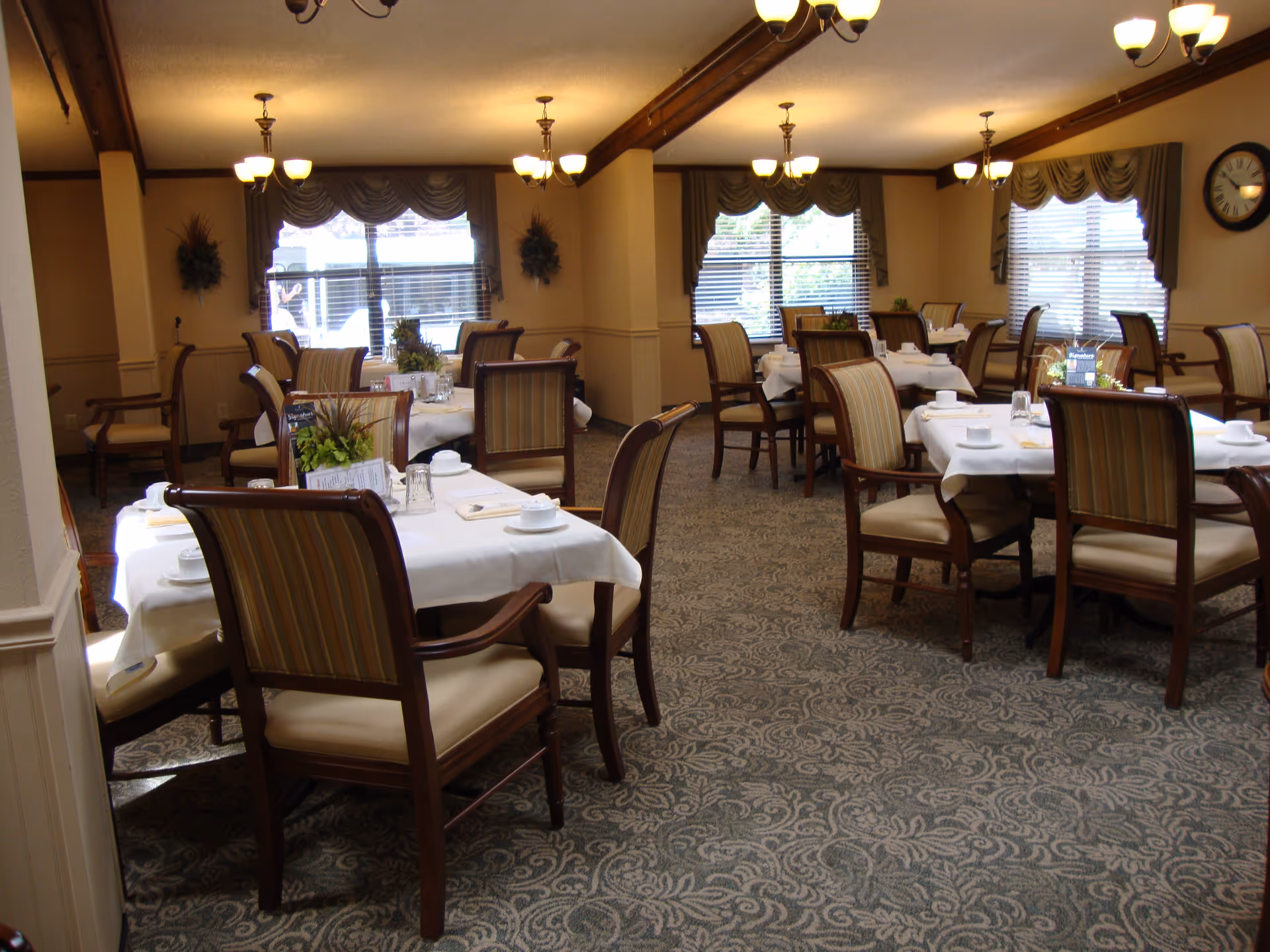 A dining room with multiple tables covered with white tablecloths, each set with cups, saucers, and glasses. The room has patterned carpet, wooden chairs with striped upholstery, large windows with blinds and valances, and ceiling light fixtures.