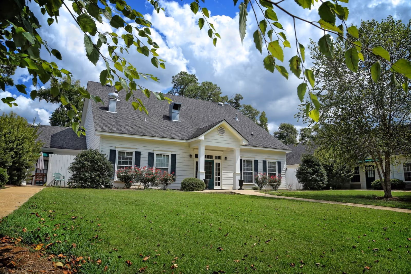 A white, single-story senior care facility with a front entrance, surrounded by green lawn and shrubs.