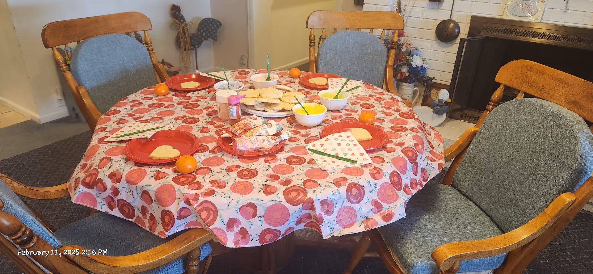 Round dining table with a floral tablecloth set with plates, bowls, snacks and four wooden chairs in a cozy room with a fireplace.