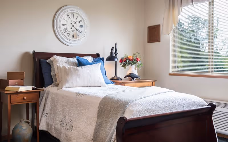 A cozy bedroom with a wooden bed frame, white and blue pillows, and a white quilted bedspread. There is a wooden nightstand on each side of the bed, one with a black lamp and a vase of flowers, and the other with a book and two small boxes. A large round clock hangs on the wall above the bed, and a window with blinds lets in natural light.