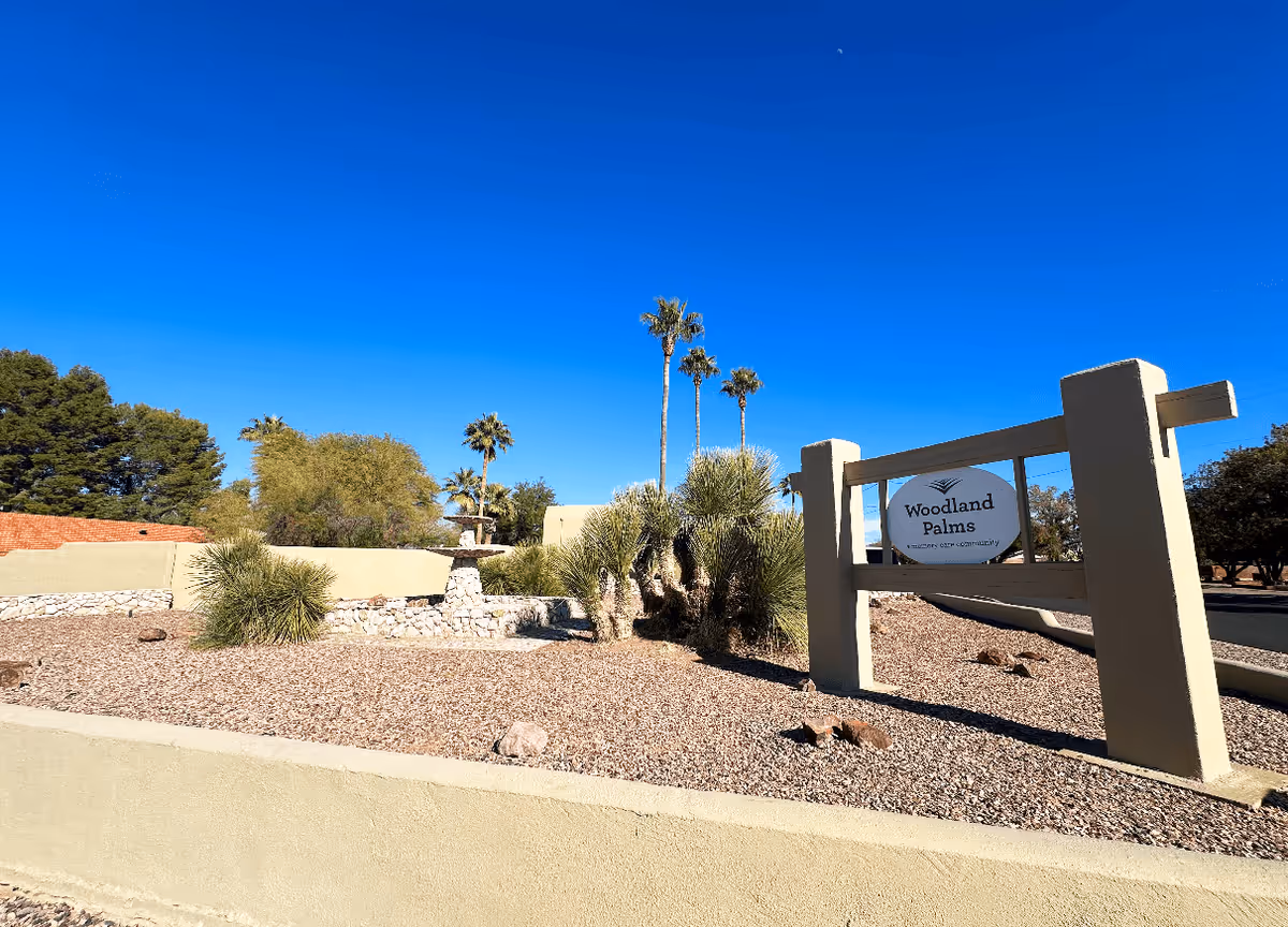 Outdoor view of the entrance area of Woodland Palms Memory Care facility featuring a sign with the facility name, desert landscaping with rocks, palm trees, and a clear blue sky.