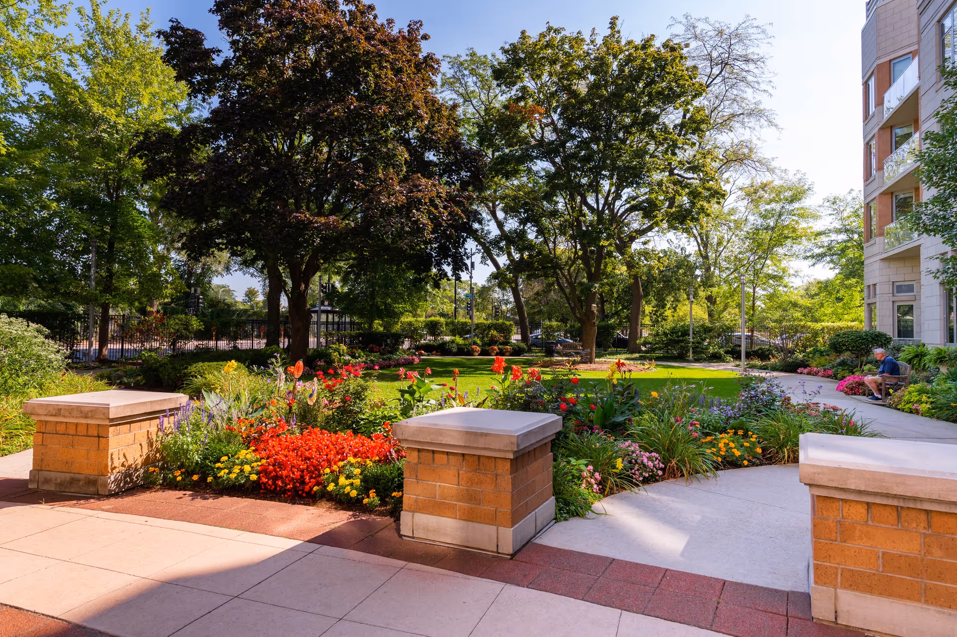 A landscaped outdoor garden area with colorful flowers, green grass, and several trees. There is a paved walkway and brick pillars in the foreground. A person is sitting on a bench near the building on the right side of the image.
