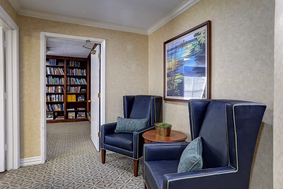 A cozy interior seating area with two dark blue wingback chairs, each with a decorative pillow, and a small round wooden table with a potted plant between them. On the wall above the chairs is a framed colorful landscape painting. An open door reveals a room with wooden bookshelves filled with books.